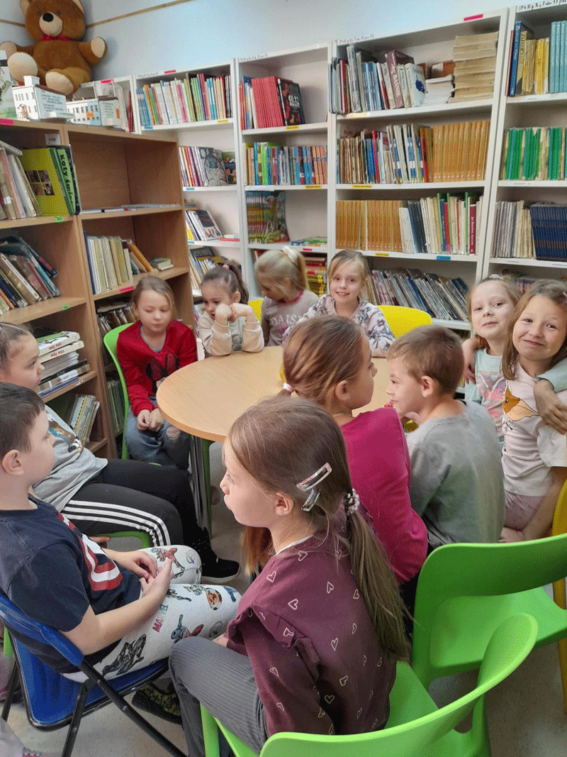 Dzieci w bibliotece siedza przy stoliku i słuchaja jak Pani z biblioteki czyta im książkę