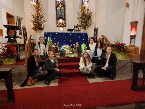 Grafika przedstawia członkinie zespołu Tirlitonki oraz Marię Dudek w kościele pw. Niepokalanego Poczęcia Najświętszej Maryi Panny w Kołobrzegu koło ołtarza przy żłóbku