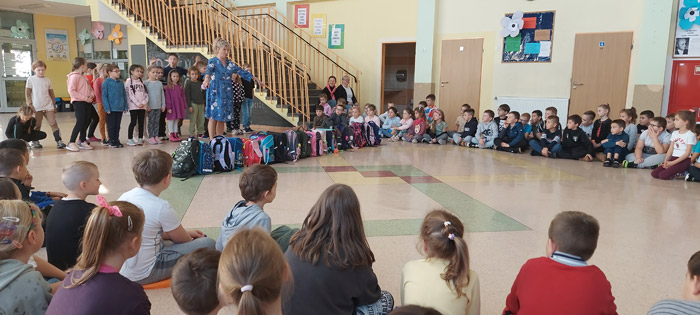 Grafika przedstawia dzieci z klasy 2a występujące na apelu wraz z wychowawcą oraz dzieci z klas 1-3 siedzące na holu