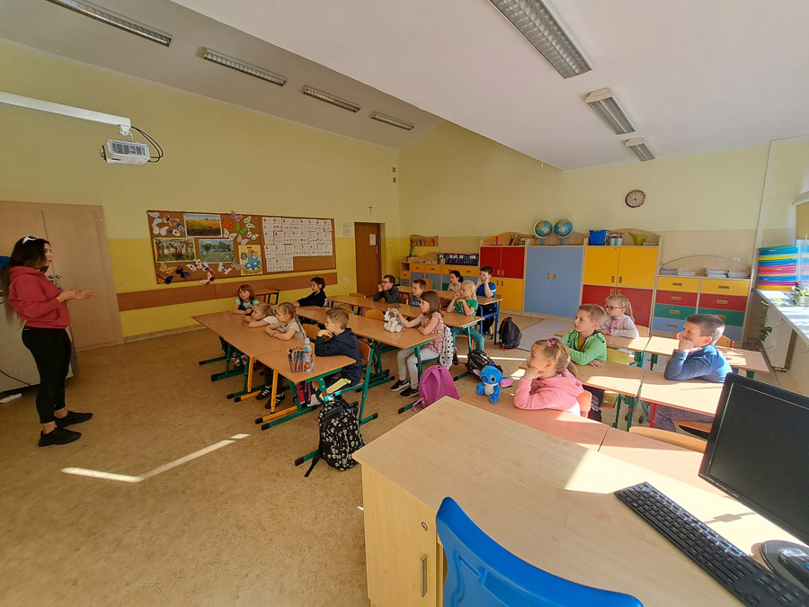 Grafika przedstawia salę lekcyjną, dzieci z klasy 1c siedzące w ławce i słuchające wypowiedzi mamy kolegi