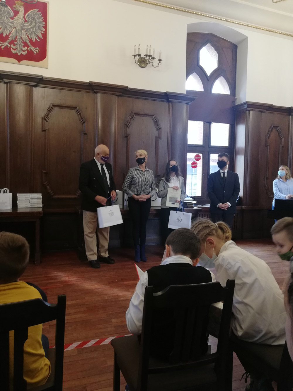 Grafika przedstawia uczniów w sali koncertowej ratusza Kołobrzeskiego oraz panią prezydent miasta
